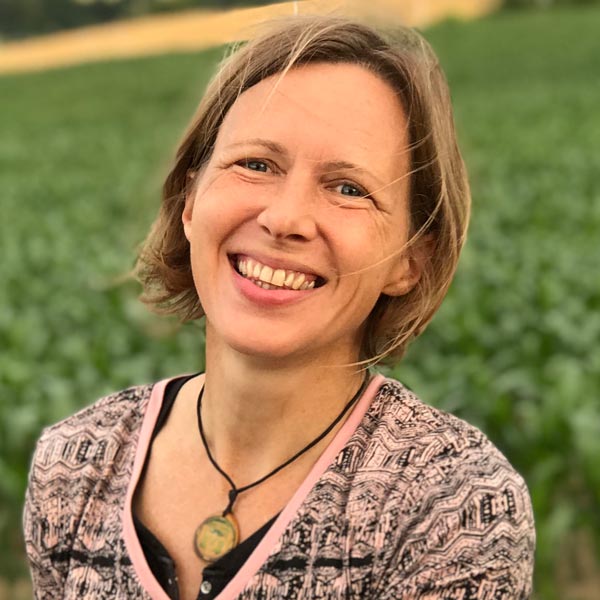 Blonde Frau sieht lächelnd in die Kamera. Im Hintergrund sind grüne Wiesen